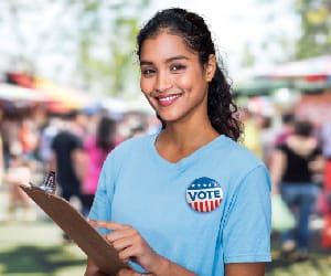 A young woman smiling and holding a clipboard with a Vote button pinned to her t-shirt.