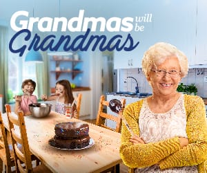 A grandmother standing at the head of a kitchen table with grandkids eating chocolate cake.