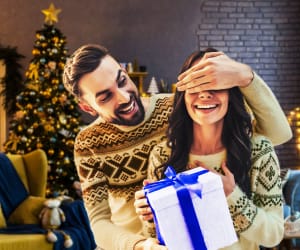 A young couple opening a holiday present in front of a christmas tree.