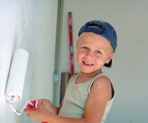 A young boy wearing a blue cap using a roller brush to paint a wall