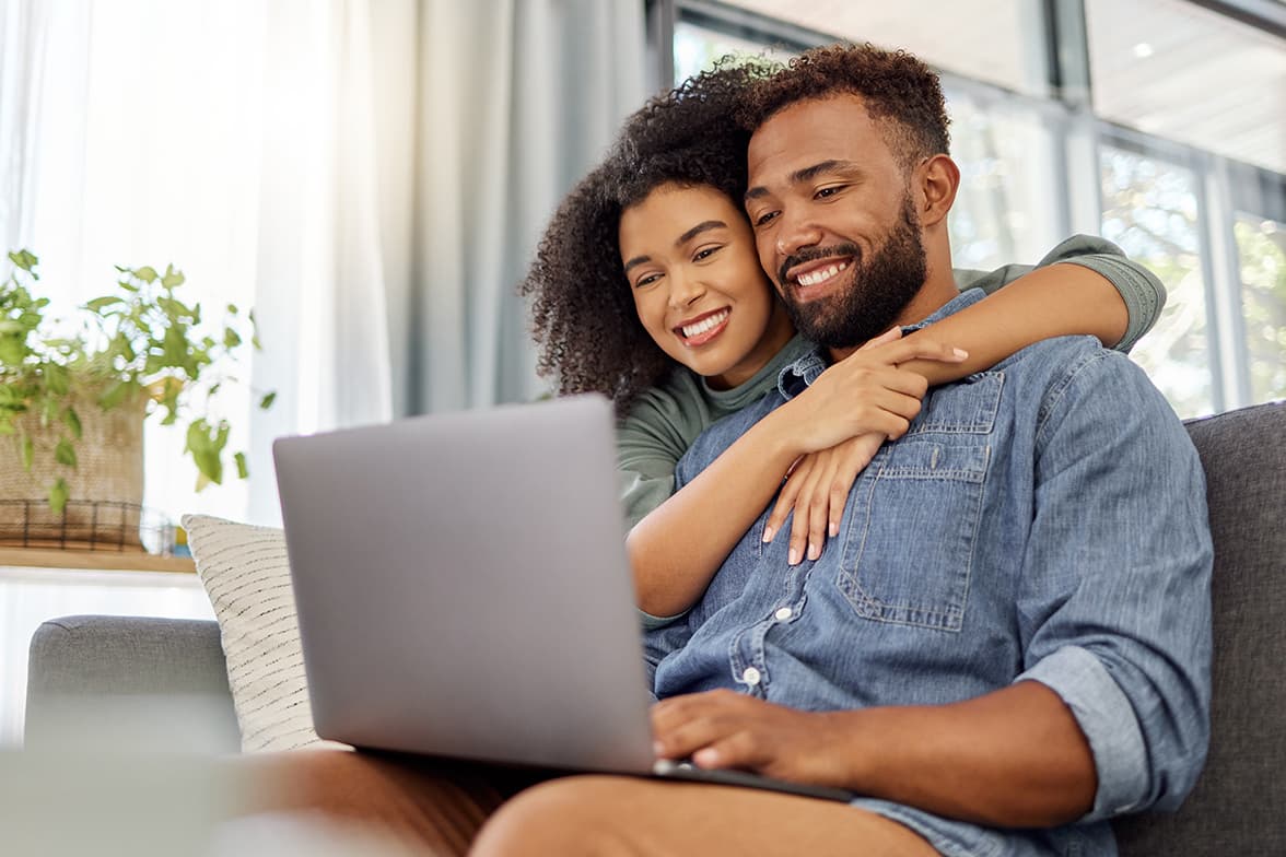 A young couple sitting on a couch looking at a laptop with excitement.