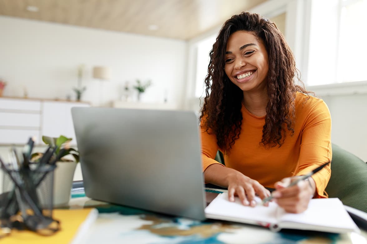 A young, smiling woman sitting at a desk with an open notebook and looking at a laptop.