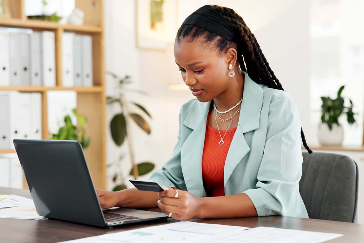A professional woman sitting at a desk using a laptop and holding a credit card.