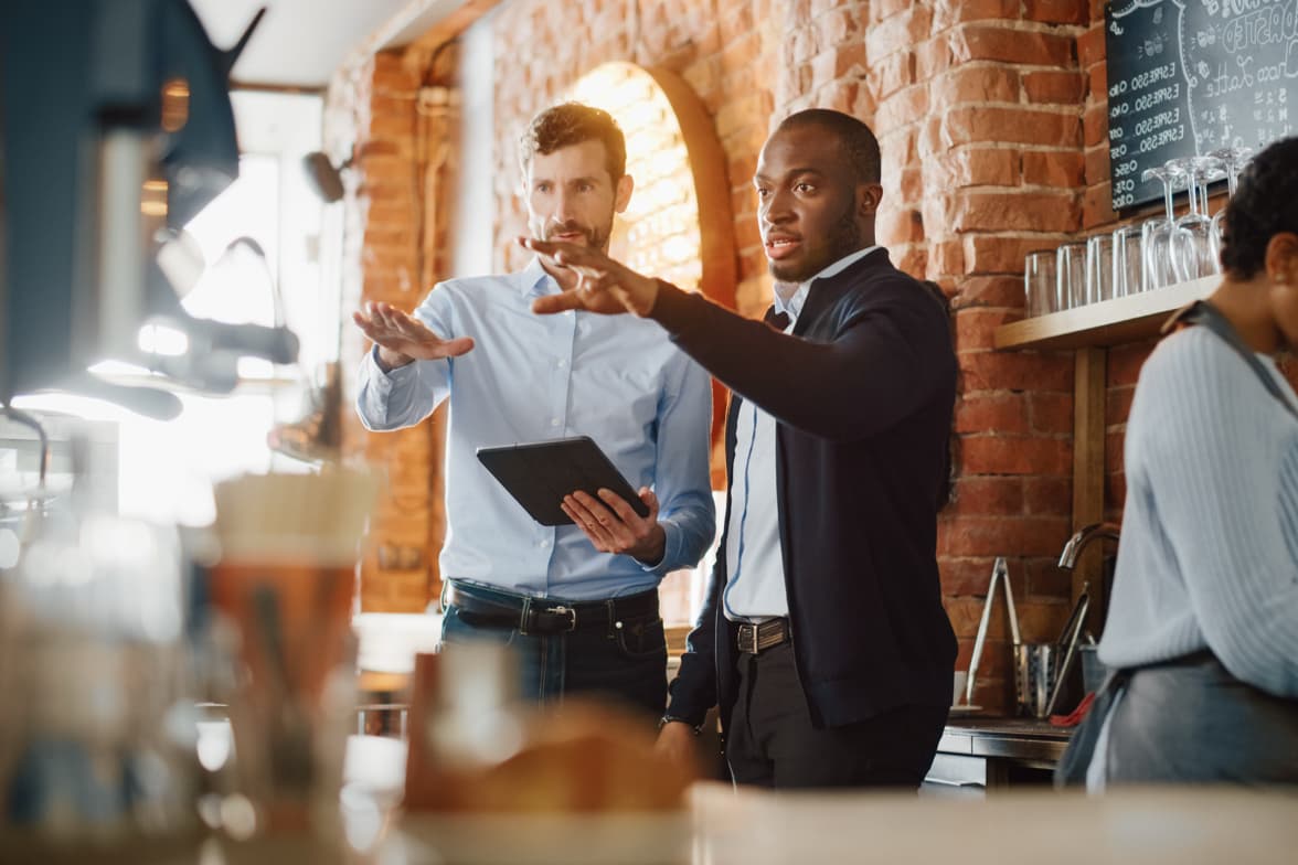 Two professonal men discuss business matters behind the counter of a coffee shop.