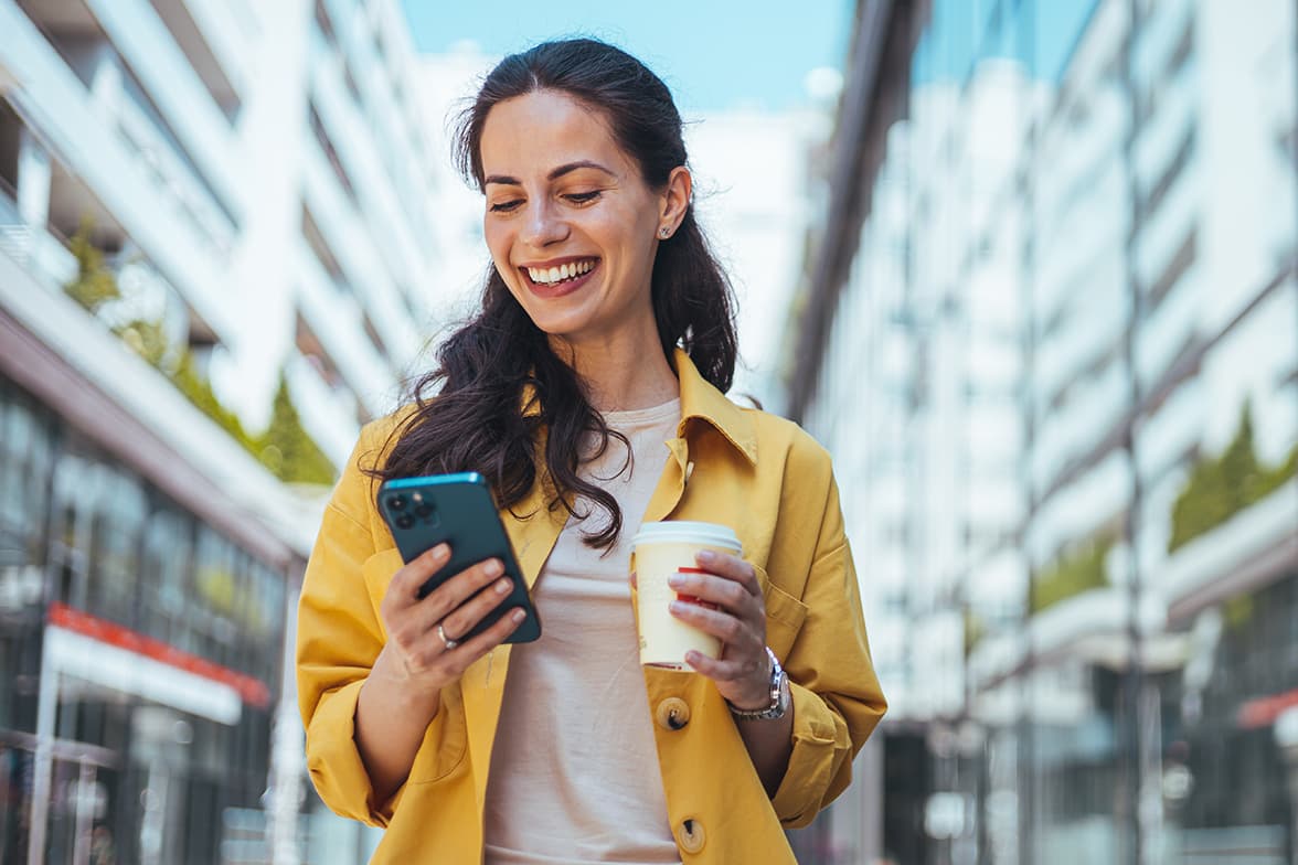 A smiling woman holding a smart phone and a coffee.