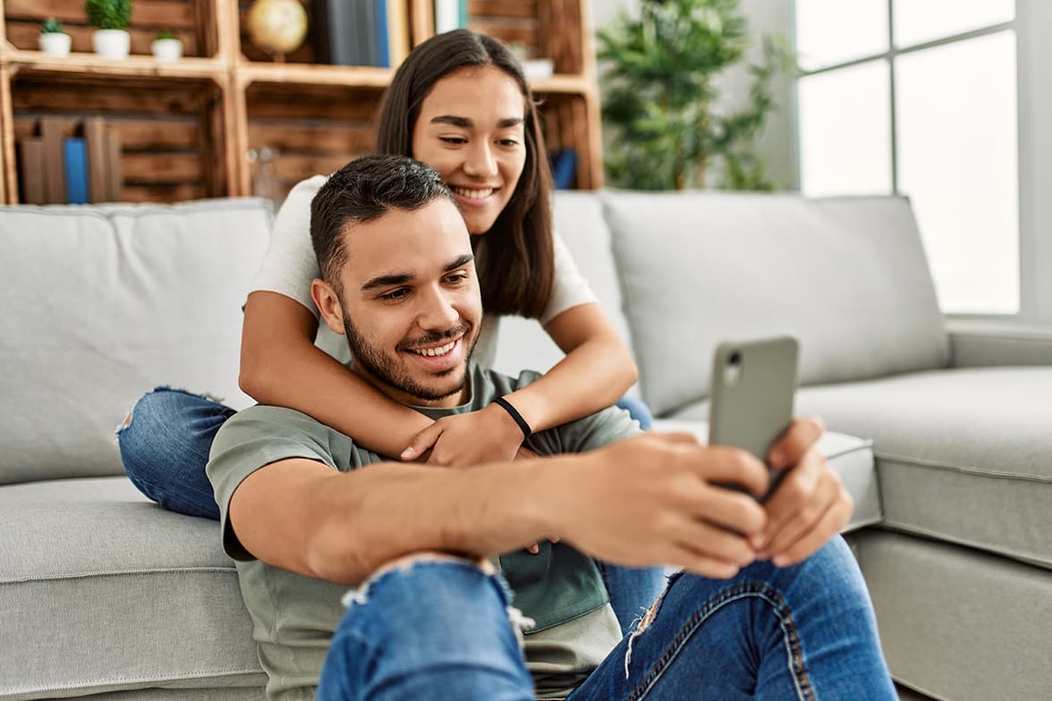A young couple sittng on a couch looking at a smart phone.