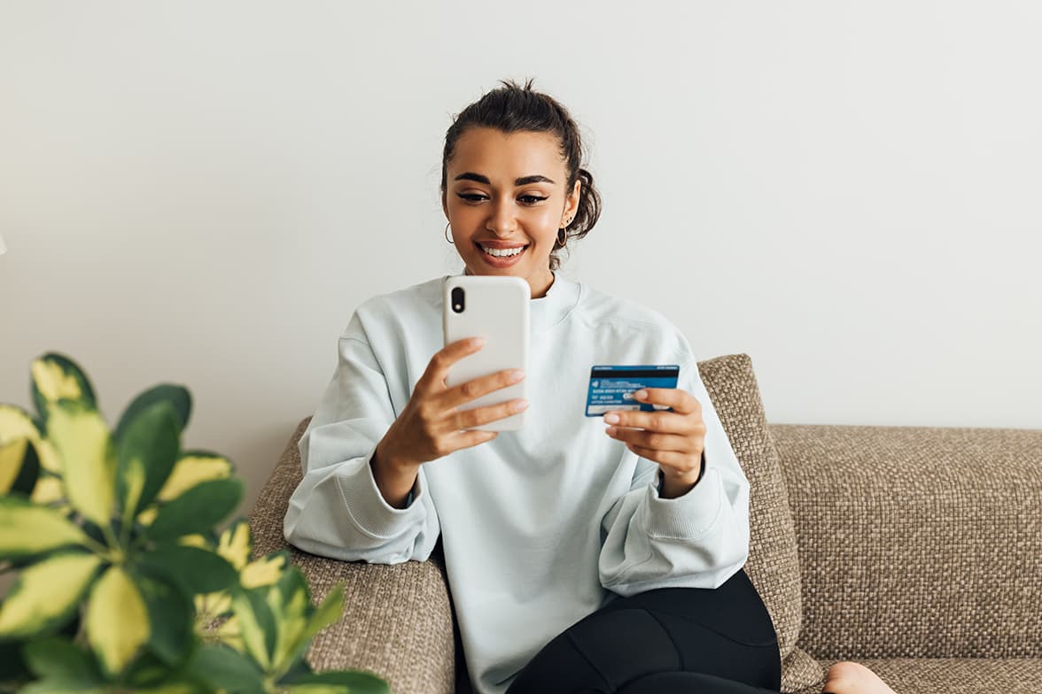 A young woman sitting on a couch holding a debit card and a smart phone.