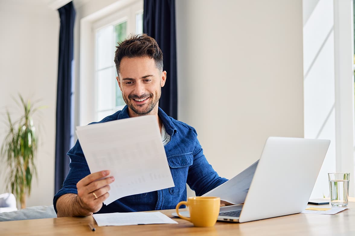 A middle-aged man sitting at a desk with a laptop open while looking at a document.