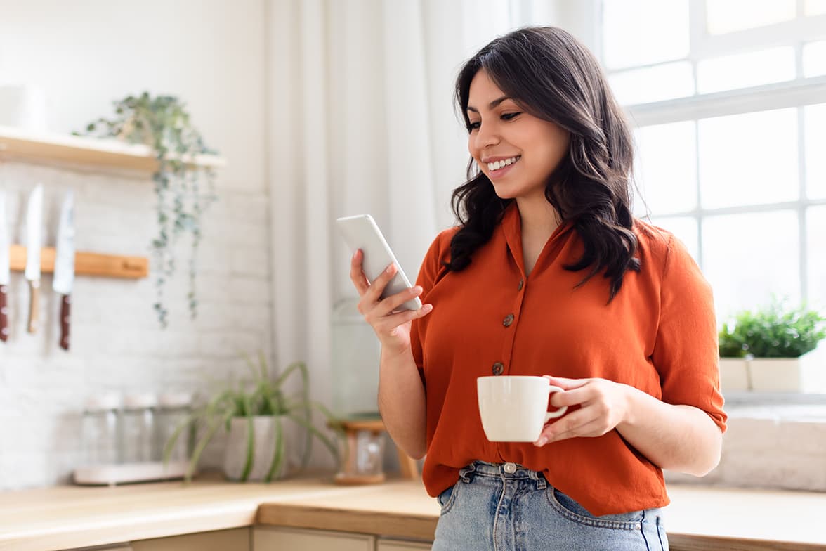 A young woman standing in a kitchen holding a mug and a mobile phone.