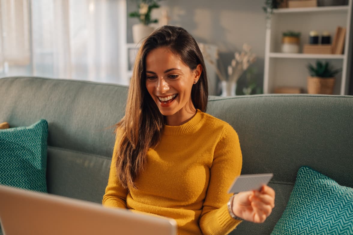 A smiling woman sitting on a couch holding a debit card while looking at a laptop.