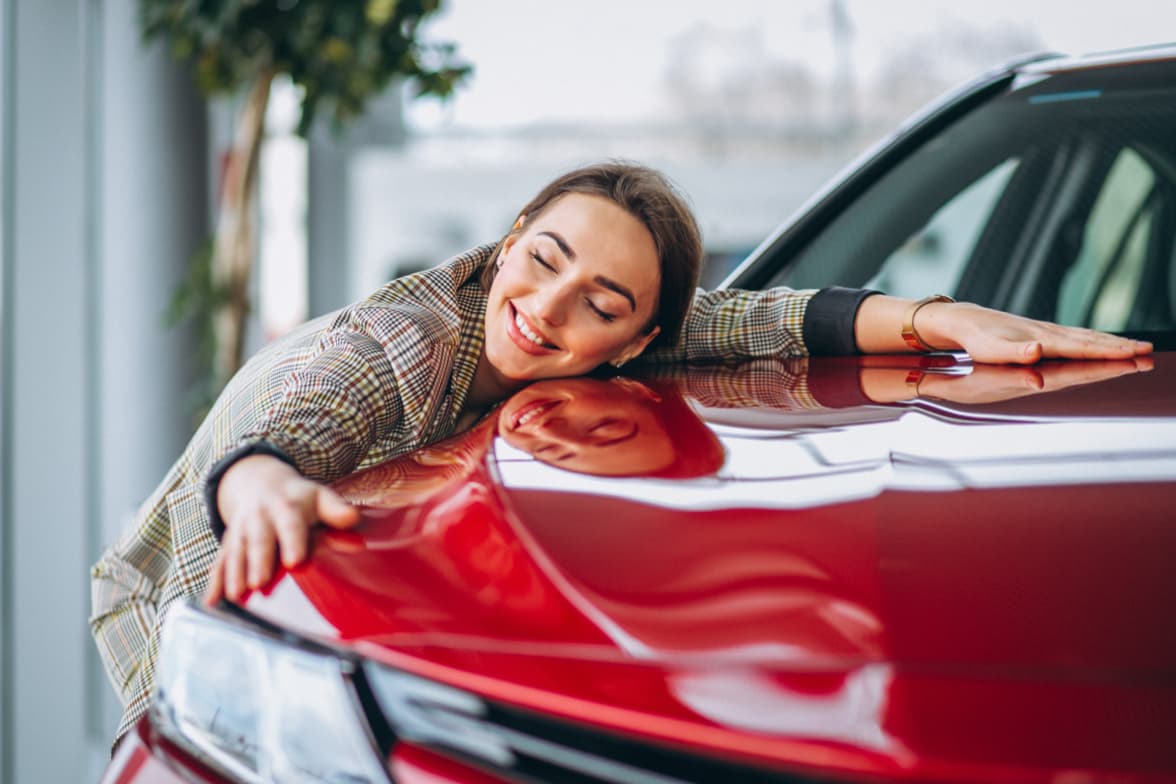 Woman smiling and embracing a red vehicle in a car dealership showroom, expressing satisfaction with a new car purchase.