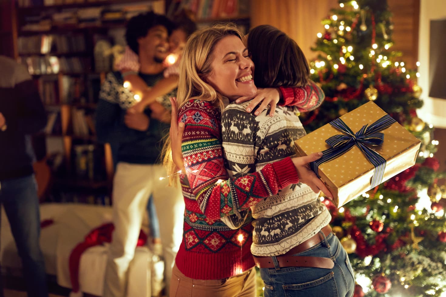 Two women hugging while holding a wrapped gift near a Christmas tree.