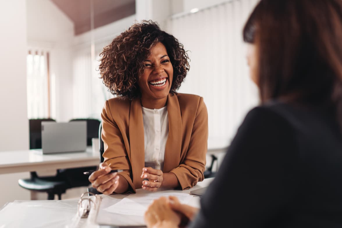 Woman smiling and meeting with another person across a desk in an office discussing documents.