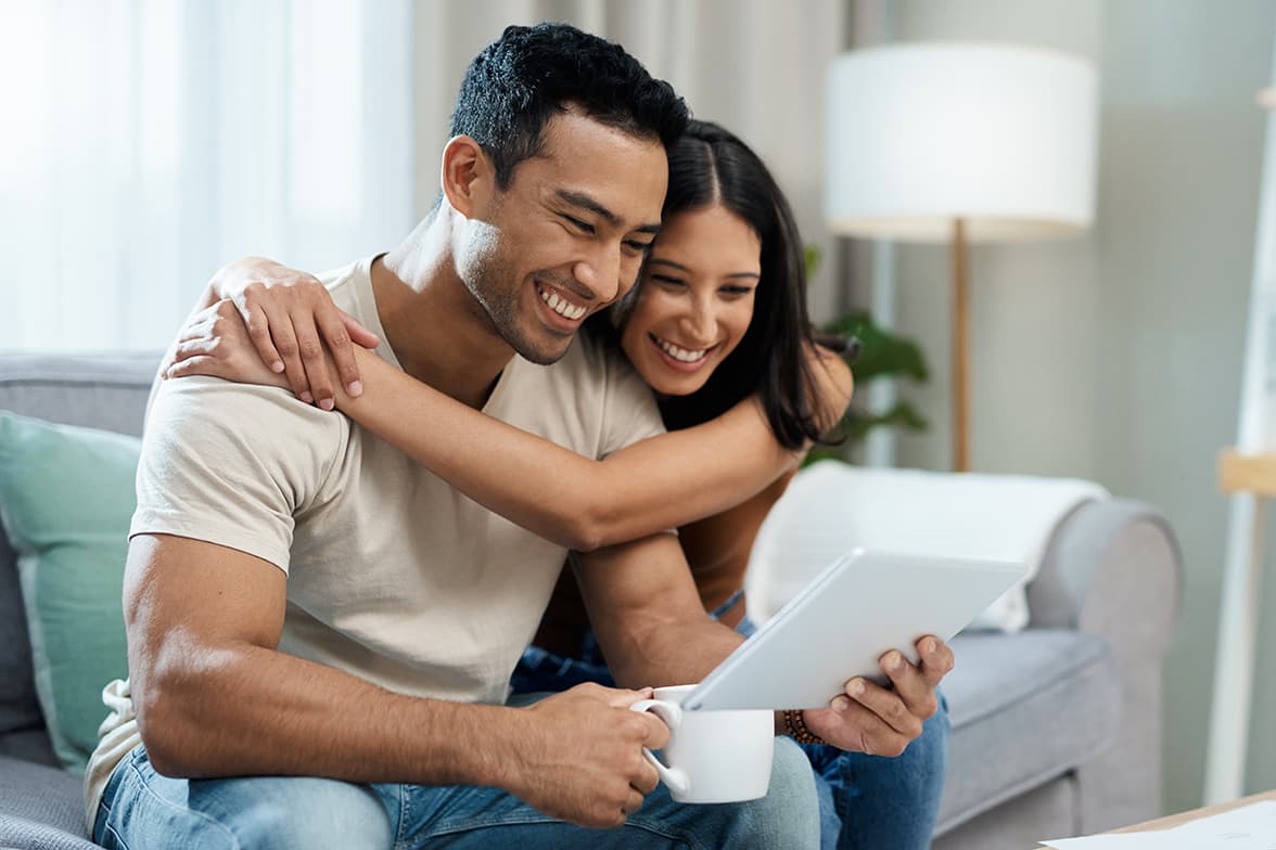 A smiling, young couple sitting on a couch looking at a tablet.