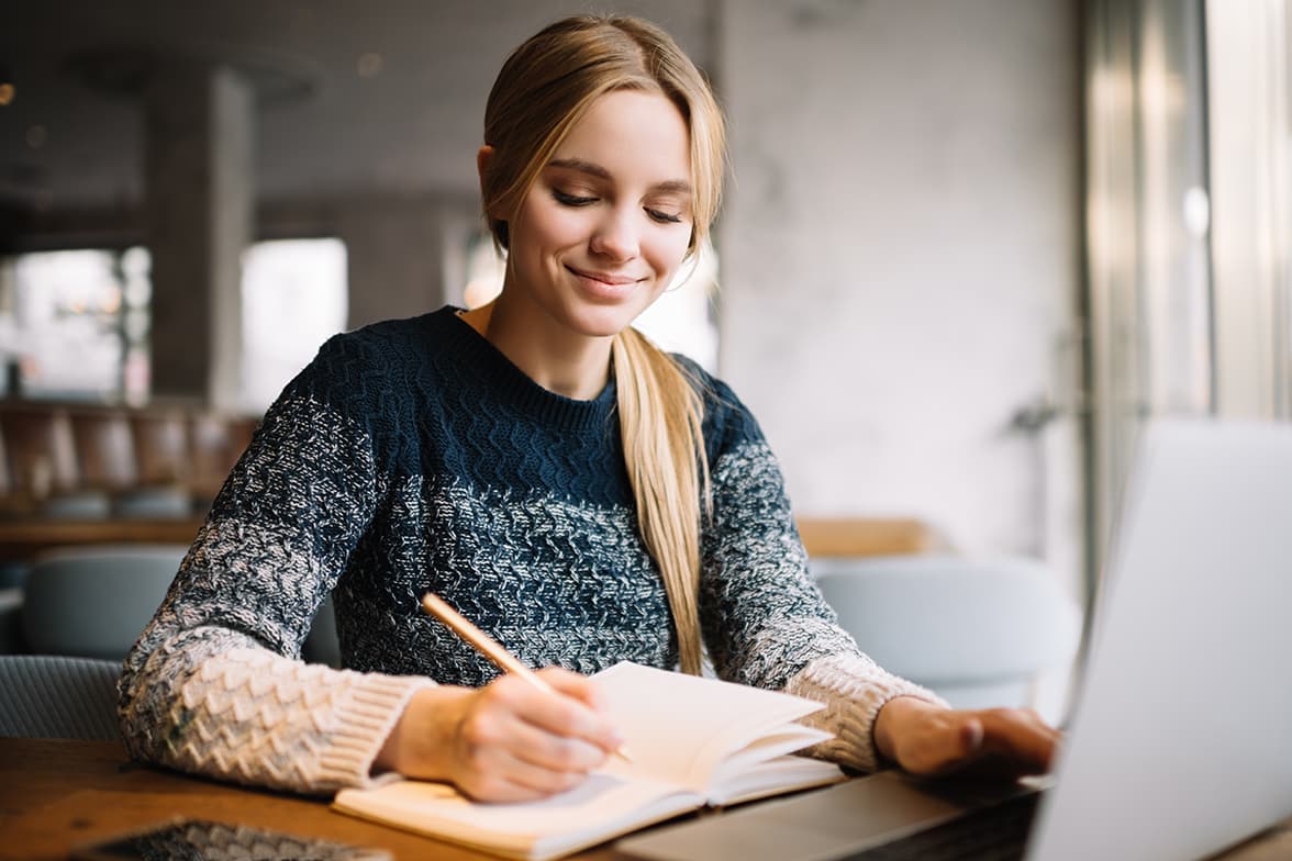 A young woman writing on a book in front of a laptop.