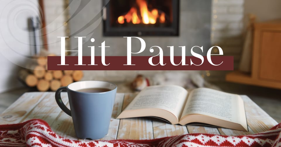 An open book resting on a wood table next to a coffee mug. In the background, a wood fireplace. Across the middle, the words: hit pause.