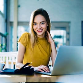 A young woman smiling while sitting at a table with an open laptop.