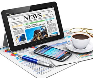 A tablet showing an online newspaper next to a coffe cup on top papers of charts and graphs